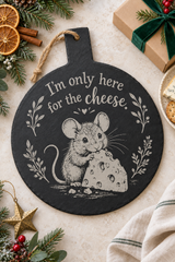A round black slate cheese board with a hanging loop, featuring a white illustration of a mouse holding cheese and the text 'I'm only here for the cheese.' It is styled in a holiday flat-lay with pine branches, dried orange slices, a wrapped gift, and a striped cloth.