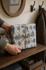 A gift box wrapped in colourful Flat Caps & Park Benches Wrapping Paper wrapping paper with a blue and purple and mustard colours on white, tied with a natural jute twine bow, held by hands on a rustic wooden shelf.