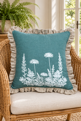 Square duck egg linen cushion with white flowers and a large natural frill resting on a wicker chair.