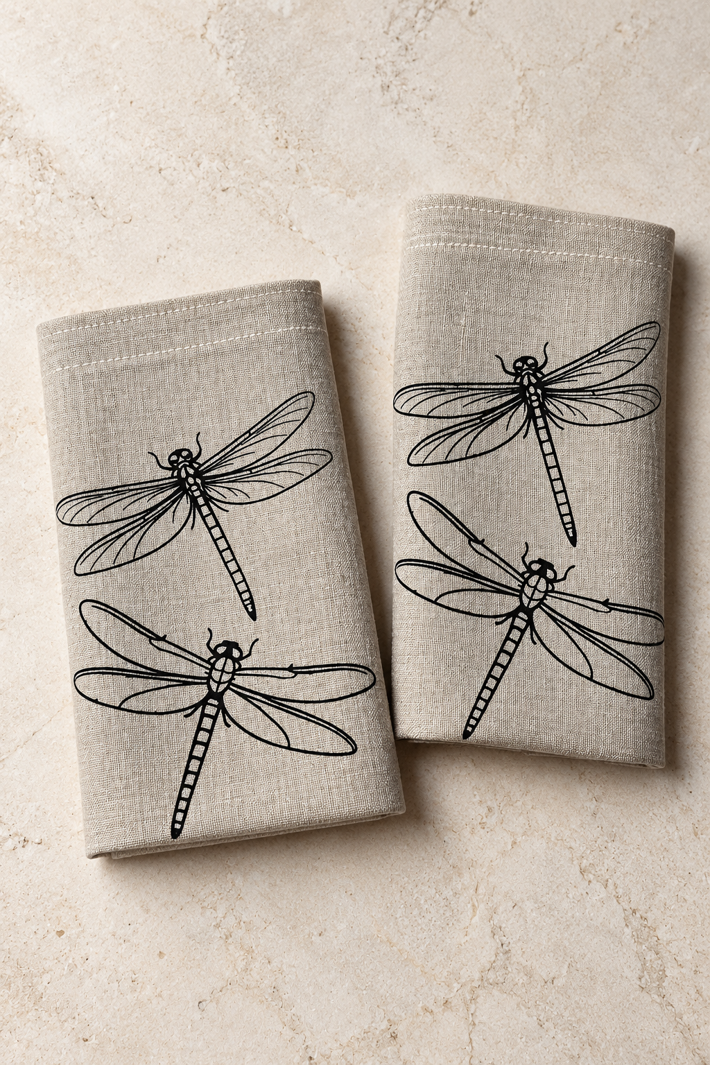 Two natural linen napkins with black dragonfly prints arranged on pale stone.