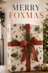Illustrated fox Christmas wrapping paper featuring a scarf-wearing fox cycling through the snow with a little dog in the front basket and a decorated fir tree and presents across the rear. The pencil-sketch design includes tartan scarves, bells, pine sprigs, baubles, parcels and tyre tracks in teal, green, mustard, red and pink on warm ivory.