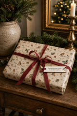 A wrapped Christmas gift box with a funny git tag with the words "this was the best i could do on Christmas eve" on it. The wrapping paper has a robin and holly pattern and a dark red ribbon, placed on a wooden table in a decorated holiday setting with pine branches, a candle, and a mirror.