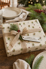 A wrapped gift box on a rustic wooden table, covered in cream wrapping paper with a green Brussels sprouts skating  holiday pattern, with a gift tag that reads "a special gift you never knew you wanted" on it all tied with a beige burlap ribbon and bow. Pine branches, red berries, plates, and a softly blurred Christmas tree lights background create a warm festive scene.