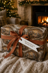 A Christmas gift box wrapped in brown and beige patterned paper with a brown ribbon and a white tag that reads 'They were out of THE GOOD STUFF', placed on a cream knit blanket in front of a cozy fireplace.