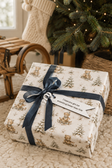 A Christmas gift box wrapped in cream paper with a teddy bear and tree pattern, tied with a navy ribbon and a white tag that reads 'I hope you get something better than this.' The box sits on a textured rug near a Christmas tree and stocking.