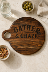 A round dark wood serving board with a handle, engraved with the words 'GATHER & GRAZE' in a rustic serif font. The board is styled in a flat-lay setting on a light beige stone surface, surrounded by a glass of water, a bowl of green olives, a bowl of nuts, and a folded beige cloth.