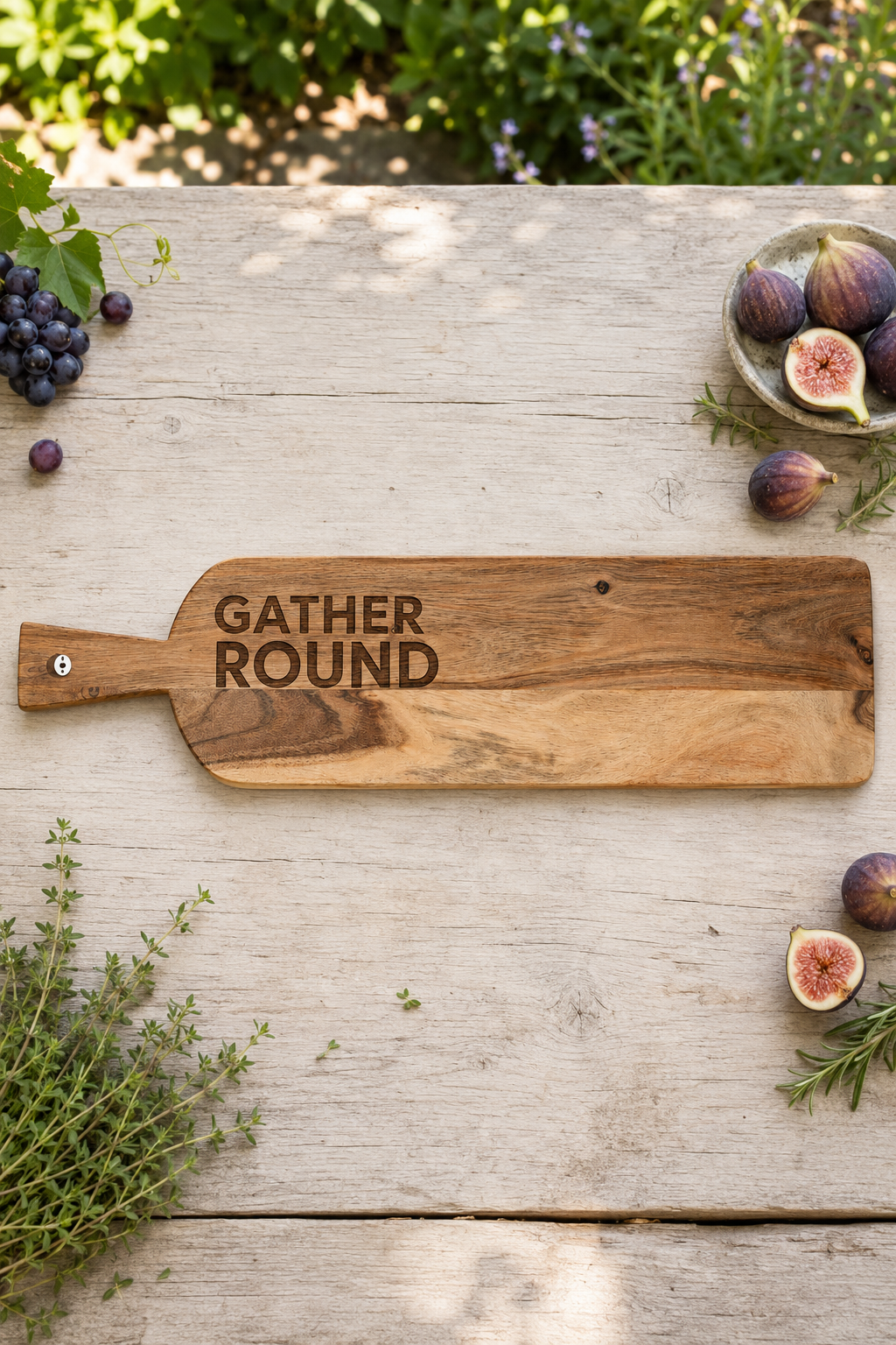 A rustic wooden serving board with the words 'GATHER ROUND' engraved on it, styled on a weathered wooden table with figs, grapes, herbs, and greenery in the background.