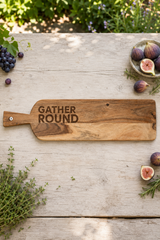 A rustic wooden serving board with the words 'GATHER ROUND' engraved on it, styled on a weathered wooden table with figs, grapes, herbs, and greenery in the background.