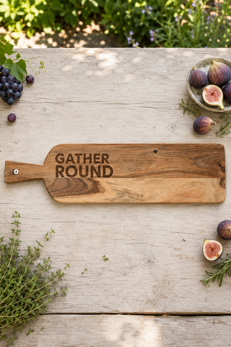 A rustic wooden serving board with the words 'GATHER ROUND' engraved on it, styled on a weathered wooden table with figs, grapes, herbs, and greenery in the background.