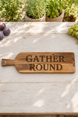 A rectangular wooden serving board with a handle, engraved with the words 'GATHER ROUND' in dark lettering, displayed on a light wooden table with grapes and potted plants in the background.