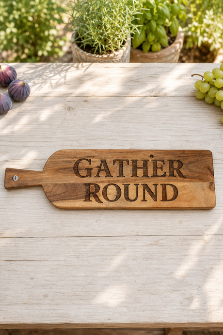 A rectangular wooden serving board with a handle, engraved with the words 'GATHER ROUND' in dark lettering, displayed on a light wooden table with grapes and potted plants in the background.