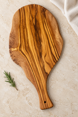 A large olive wood serving board with a decorative handle, shown from a top-down view on a textured beige stone surface with a sprig of rosemary and a folded cream cloth.