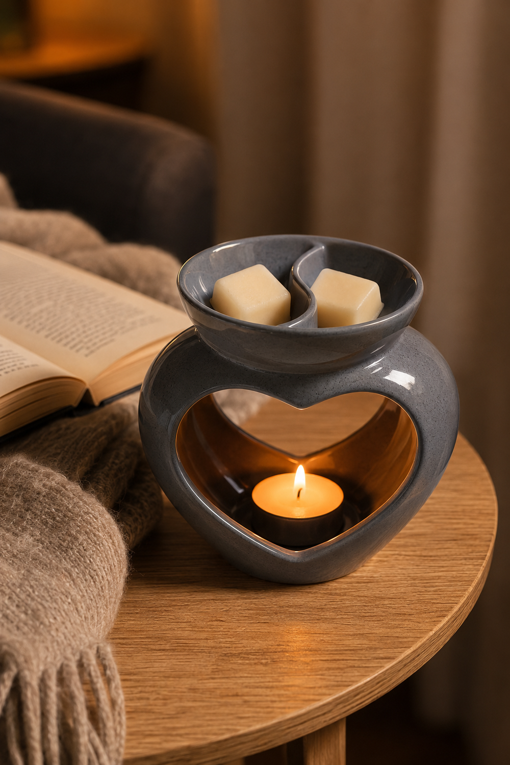 A gray ceramic heart-shaped candle warmer with a lit tea light candle inside, shown on a wooden table next to an open book and a beige knit blanket.
