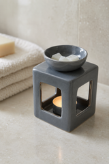A dark gray ceramic cube candle holder with a matching bowl on top containing white wax cubes and a glowing tea light candle inside, set on a beige stone surface with folded towels in the background.