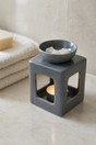 A dark gray ceramic cube candle holder with a matching bowl on top containing white wax cubes and a glowing tea light candle inside, set on a beige stone surface with folded towels in the background.