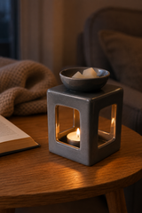 A square gray ceramic candle holder with a lit tea light candle inside and a small bowl on top holding white wax melts, placed on a wooden table in a cozy indoor setting.