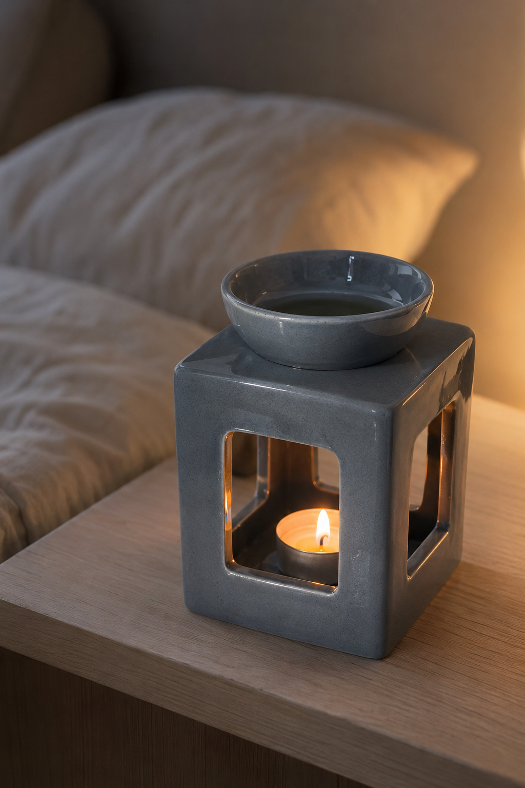 A gray ceramic cube-shaped candle warmer with a small matching bowl on top, shown lit on a wooden bedside table with a pillow in the background.
