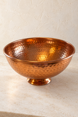 Hammered copper finish bowl with a smooth rim and low round pedestal on a pale stone surface.