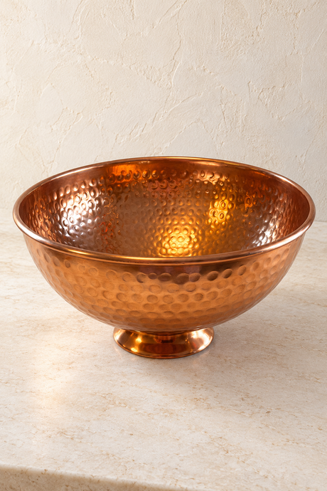 Hammered copper finish bowl with a smooth rim and low round pedestal on a pale stone surface.