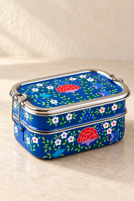 A rectangular blue metal tiffin style lunch box with a floral and mushroom pattern, featuring a silver metal clasp lid, set on a beige textured surface.