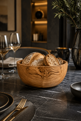 AHand-Turned Wooden Snack & Serving Bowl – 16cm filled with rustic bread slices, set on a dark stone dining table with wine glasses, plates, and a vase in a softly lit interior.