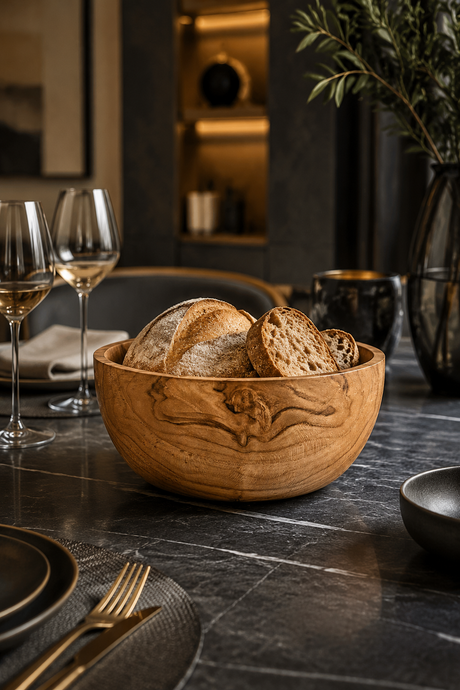 AHand-Turned Wooden Snack & Serving Bowl – 16cm filled with rustic bread slices, set on a dark stone dining table with wine glasses, plates, and a vase in a softly lit interior.