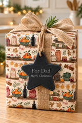 A Christmas-themed gift box wrapped in a warm-toned holiday print with illustrations of gingerbread cookies, oranges, bottles, and cinnamon sticks, tied with a burlap ribbon and bow. A slate star-shaped gift tag hangs from the front and reads 'For Dad Merry Christmas.