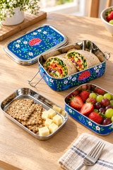 A set of decorative stainless steel lunch boxes arranged on a wooden table, shown open with a wrap, crackers, cheese cubes, and a fruit tray. The lunch boxes have a blue floral pattern with red flowers and white flowers, and are styled with a napkin and fork.