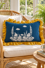 Rectangular blue botanical linen cushion with mustard ruffle on a wicker chair beside a fern.