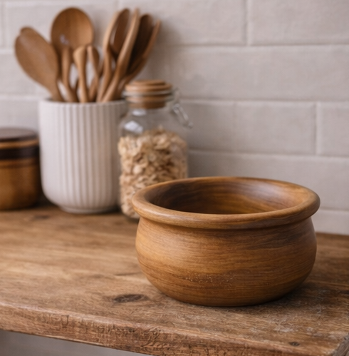 Wooden bowl on a wooden surface with kitchen utensils and jars in the background.