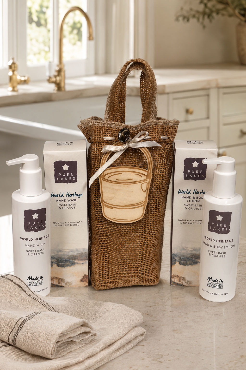 A gift set of World Heritage hand wash and body wash products arranged on a bathroom counter with a burlap tote bag, two white pump bottles, two product boxes, and a folded towel.