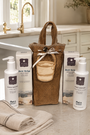 A gift set of World Heritage hand wash and body wash products arranged on a bathroom counter with a burlap tote bag, two white pump bottles, two product boxes, and a folded towel.