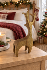 A rustic wooden reindeer figurine stands on a light wood table in a cozy, decorated bedroom setting with a bed, holiday garland, and Christmas tree in the background.