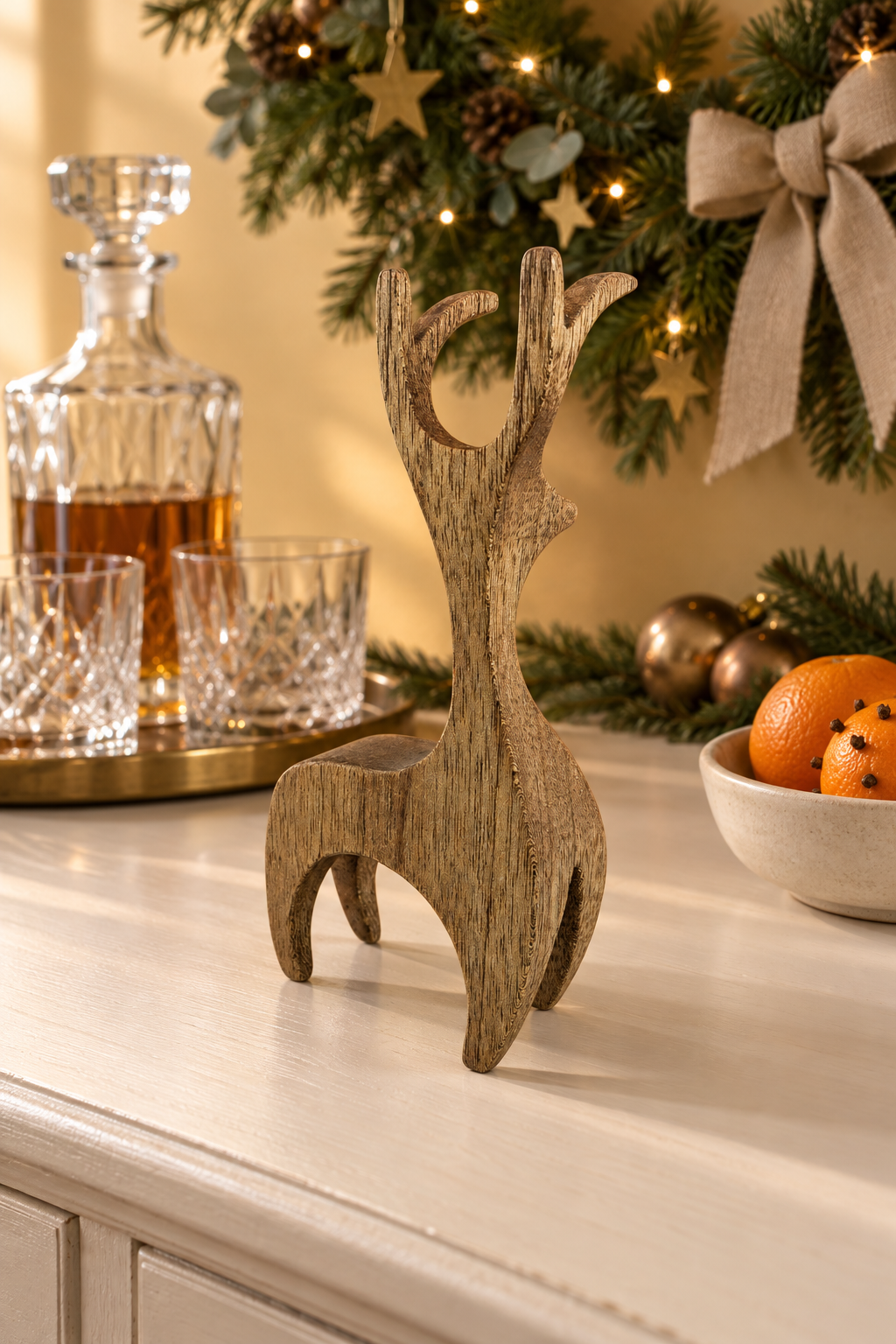 A small wooden stag figurine stands on a white cabinet in a warm holiday setting, with a glass decanter, crystal glasses, oranges, and a decorated Christmas tree in the background.