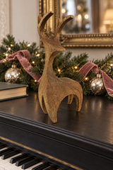 A rustic wooden reindeer figurine with antlers stands on a black piano lid, surrounded by Christmas greenery, velvet ribbons, and silver ornaments in a warm indoor setting.