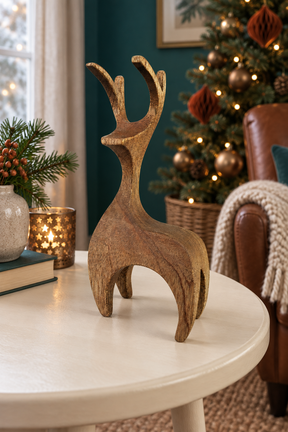 A rustic wooden reindeer figurine with a weathered brown finish stands on a round white table in a cozy, decorated living room with a Christmas tree in the background.