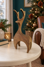 A rustic wooden reindeer figurine with a weathered brown finish stands on a round white table in a cozy, decorated living room with a Christmas tree in the background.
