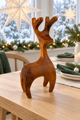 A carved wooden reindeer figurine with natural wood grain stands on a light wood dining table in a decorated holiday setting with a window, star lights, and festive greenery.