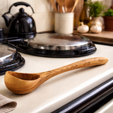Wooden ladle on a kitchen counter with a stove and various kitchen items in the background.