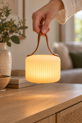 Hand holding the brown carry strap of a glowing Mochi Pure portable lamp above a console table.
