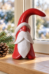 A small red and white Santa figurine with a curved hat and long white beard, displayed on a wooden windowsill with a pinecone and evergreen branches nearby.
