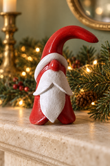 A small painted Santa holiday decoration with a red hat, red body, and white beard, standing on a stone mantel with Christmas greenery and warm string lights in the background.