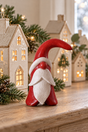A small red and white Santa figurine stands on a wooden surface in front of decorative white house ornaments with warm lights and green pine branches.
