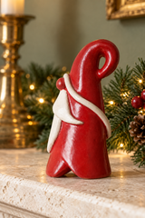 A small red Santa-like Santa figurine with a white beard and white scarf detail, displayed on a stone mantel with holiday greenery and warm lights in the background.
