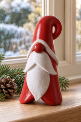 A small red and white Santa figurine standing on a wooden windowsill, with pine branches and a pinecone nearby and a snowy outdoor scene visible in the background.