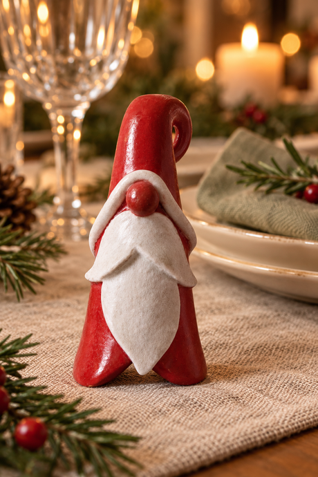 A small red and white gnome Santa on a table runner in a warmly lit holiday table setting with candles, pine branches, and plates in the background.