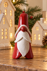 A small Christmas Santa figurine with a glossy red hat and coat, white beard, and red nose, standing on a wooden surface with miniature houses and a Christmas tree in the background.