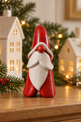 A small red and white Santa-style Santa figurine stands on a wooden surface, with decorative white house lanterns, evergreen branches, and warm string lights blurred in the background.