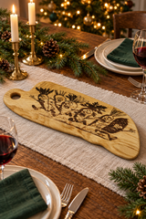 A wooden serving board with engraved vegetable cartoon characters, on a decorated holiday table setting with candles, pine branches, plates, wine glasses, and a blurred Christmas tree in the background.