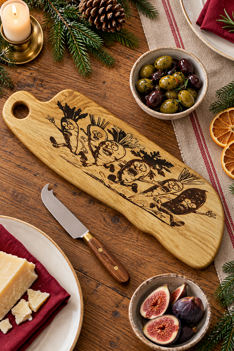 A wooden chopping board with engraved winter vegetables, styled on a rustic wooden table with cheese, figs, olives, a knife, and holiday decorations.